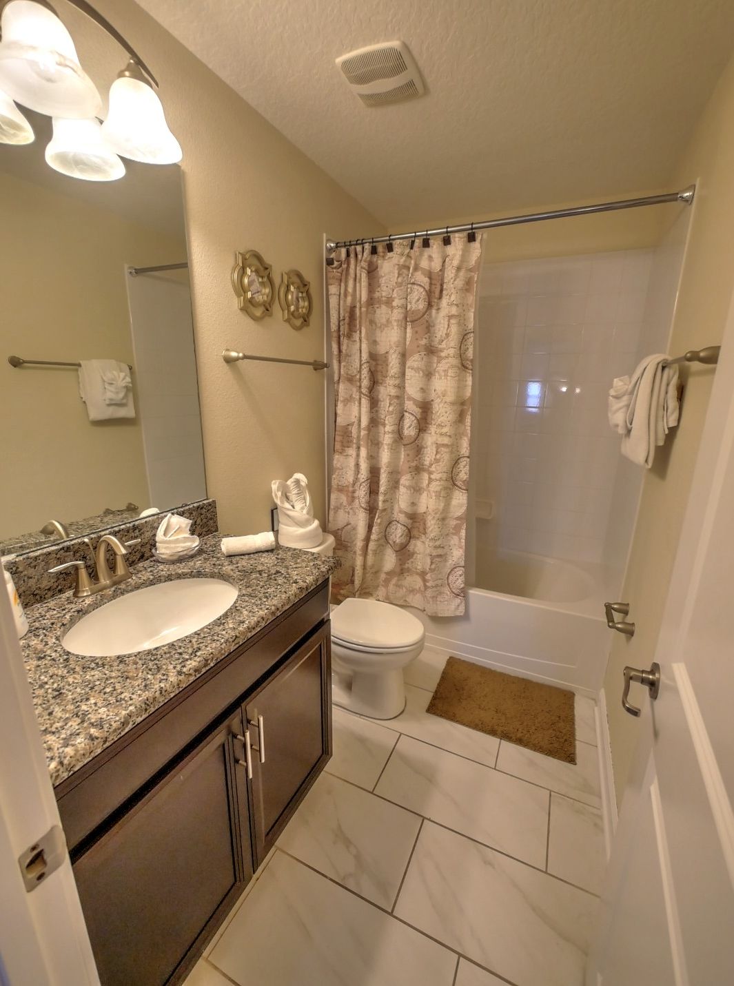 A bathroom with a granite countertop sink, a toilet, a shower with a patterned curtain, and a mirror with lights.