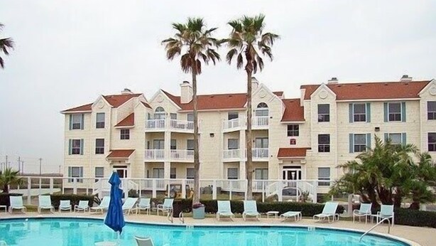 A swimming pool with lounge chairs and palm trees, surrounded by a multi-story building with balconies.