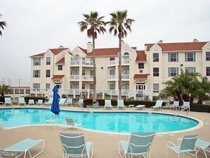 Una piscina con sillas de descanso y palmeras, rodeada por un edificio de varios pisos con balcones.