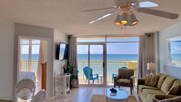 A spacious living room with a ceiling fan, a TV mounted on the wall, a balcony with a view of the ocean, and a seating area with a couch and chairs.