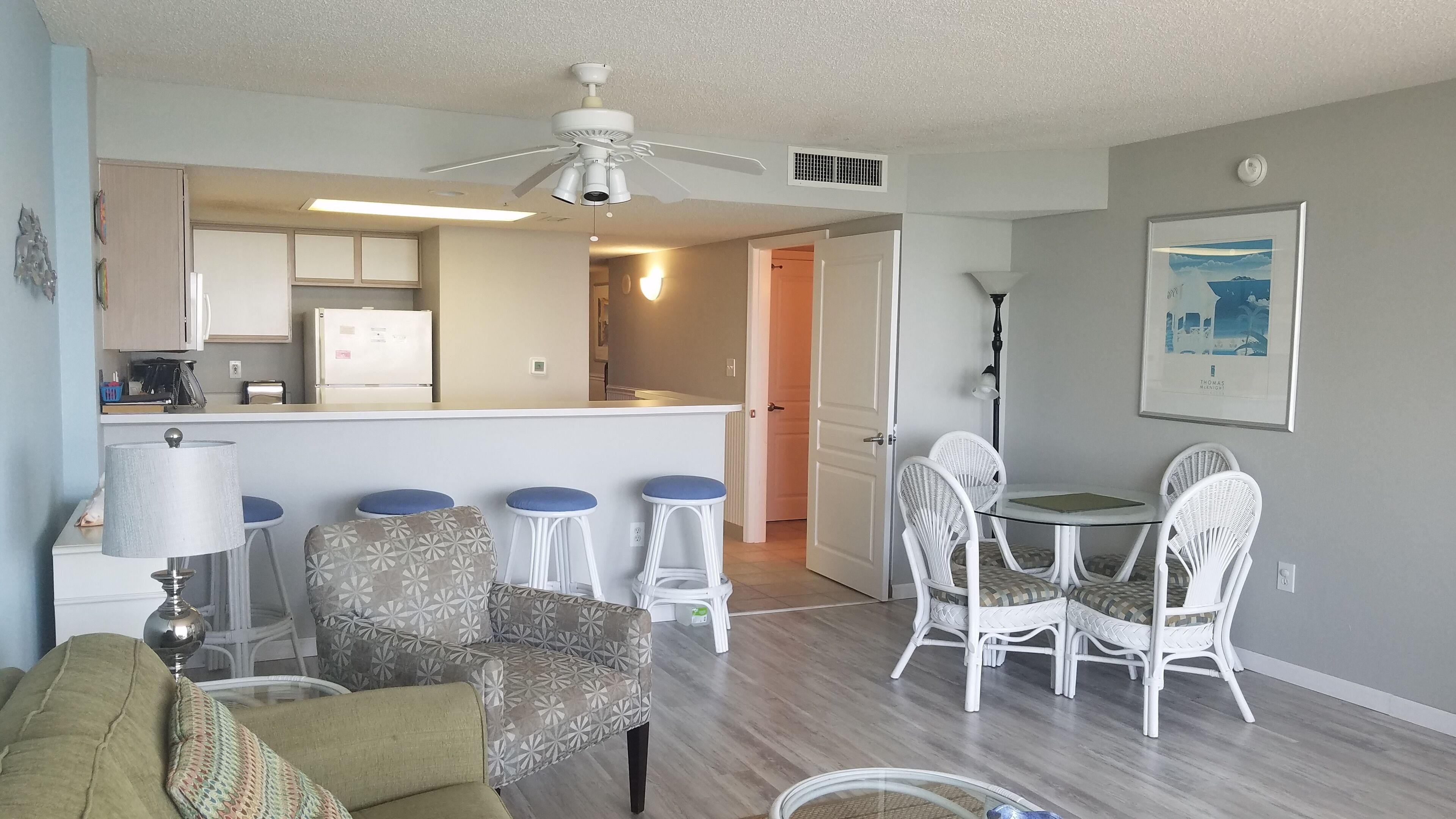 A living room with a sofa, a dining table with chairs, a kitchen area with a refrigerator, and a ceiling fan.