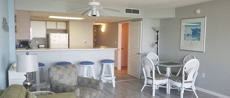 A living room with a sofa, a dining table with chairs, a kitchen area with a refrigerator, and a ceiling fan.