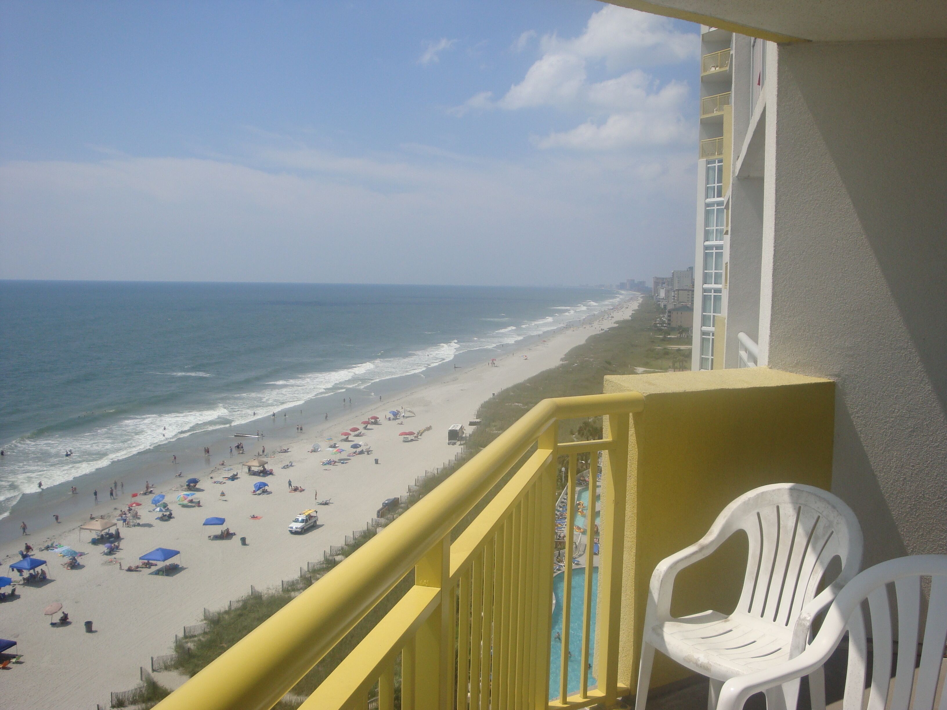 A balcony with a view of a beach, white plastic chairs, and a yellow railing.