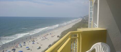 A balcony with a view of a beach, white plastic chairs, and a yellow railing.