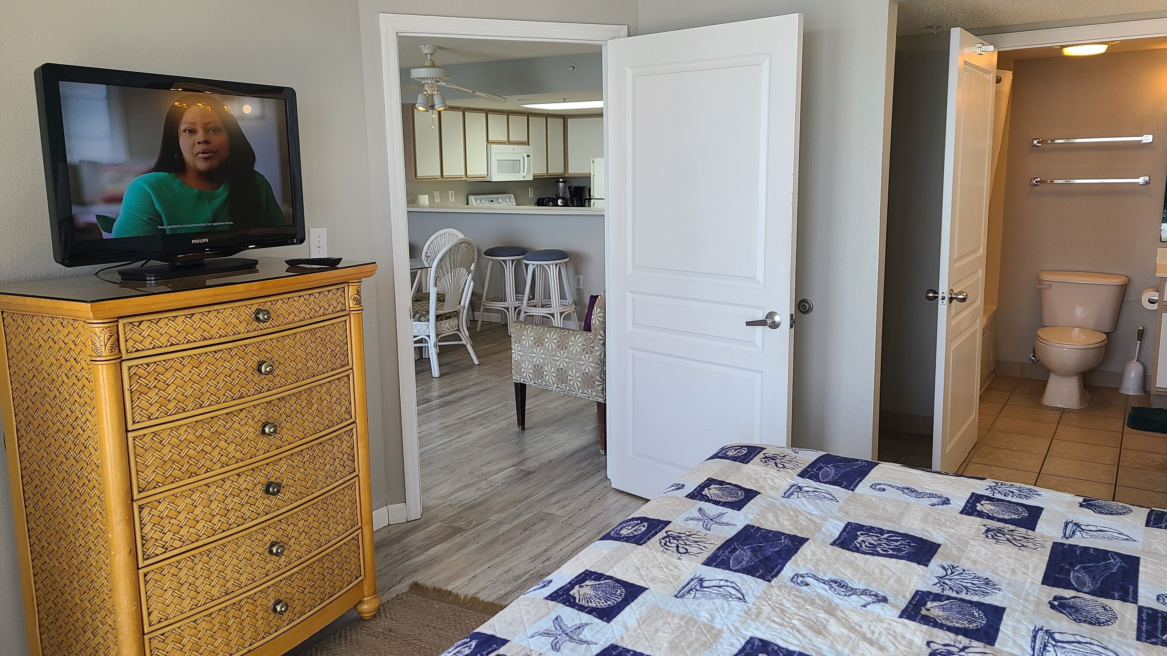 Ein Schlafzimmer mit Fernseher auf einer Kommode, einem Bett mit gemustertem Überwurf und einer offenen Tür zu einer Küche und einem Badezimmer.