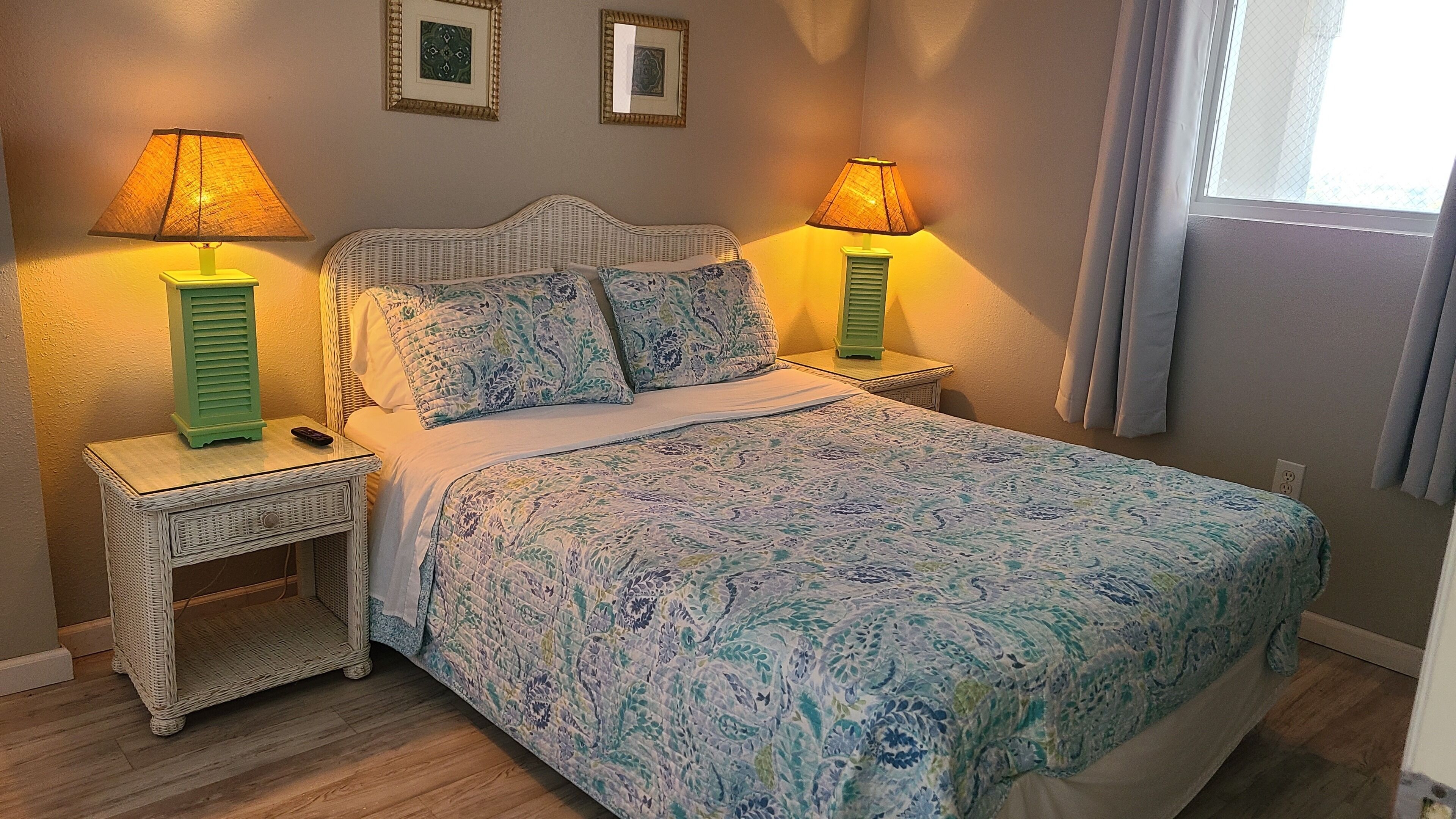 Ein ordentlich bezogenes Bett mit einem blumigen Überwurf, zwei Nachttischlampen und einem Korbnachttisch mit Fernbedienung darauf.