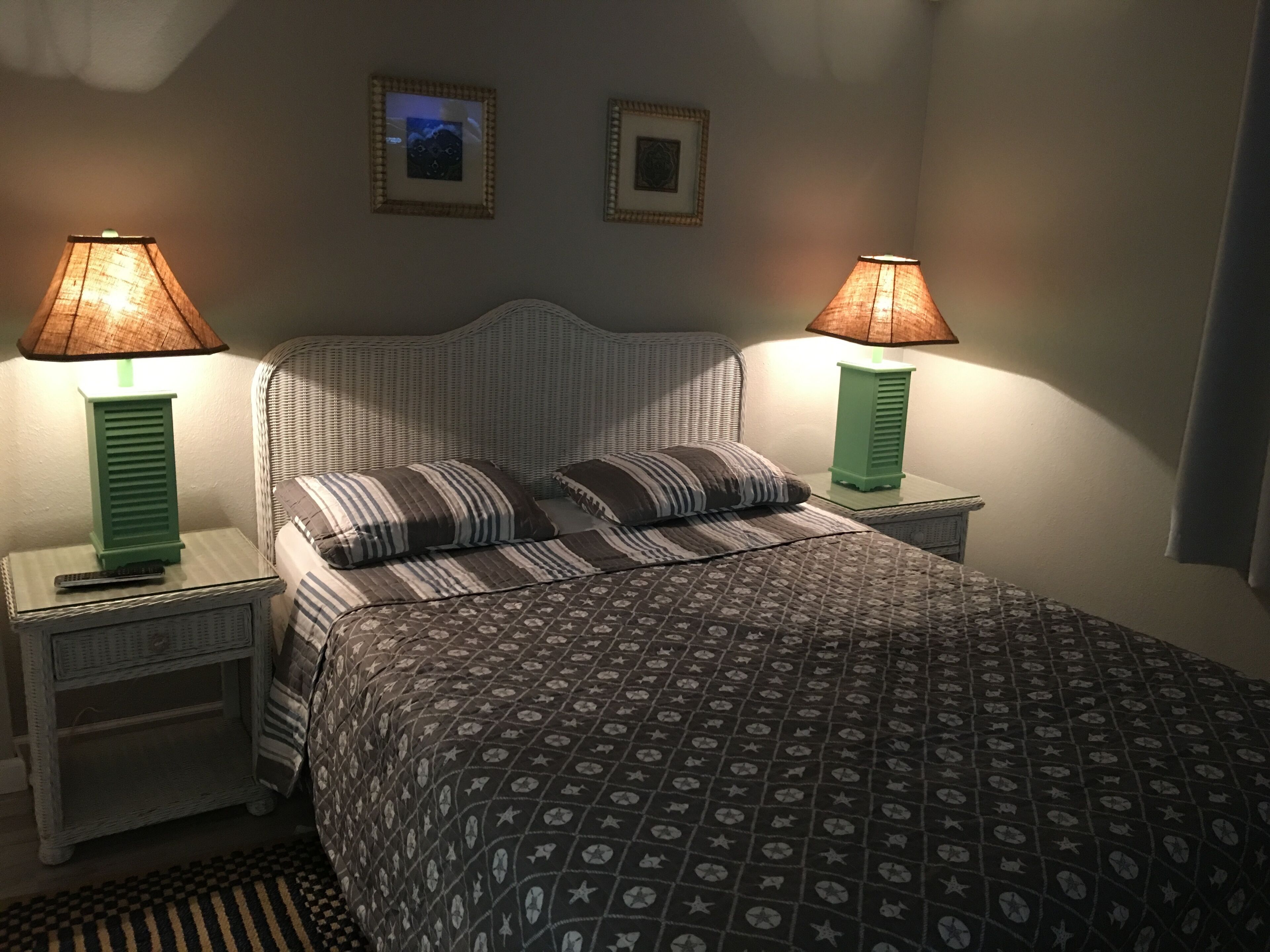 Ein Schlafzimmer mit einem gemusterten Bettbezug, zwei Nachttischlampen, einem Rattan-Kopfstück und zwei gerahmten Bildern an der Wand.
