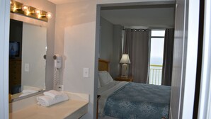 A hotel room with a bathroom, a bed with a blue patterned bedspread, a nightstand with a lamp, and a view of the balcony.