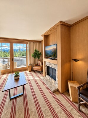 A room with a fireplace, a television, a sofa, a chair, and a view of mountains through large windows.