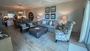 A spacious living room with a grey sofa, patterned armchair, and ottoman. There is a ceiling fan, a kitchen in the background, and framed pictures on the wall.