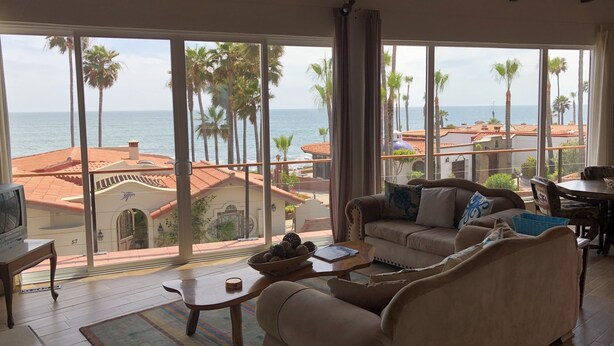 Un salon spacieux doté d’une grande fenêtre offrant une vue imprenable sur la côte, avec un canapé, une table basse et une vue sur les palmiers et la mer.
