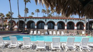 Un espace piscine avec des chaises longues blanches et un bâtiment doté de fenêtres en arc de cercle et d’un toit recouvert de tuiles.