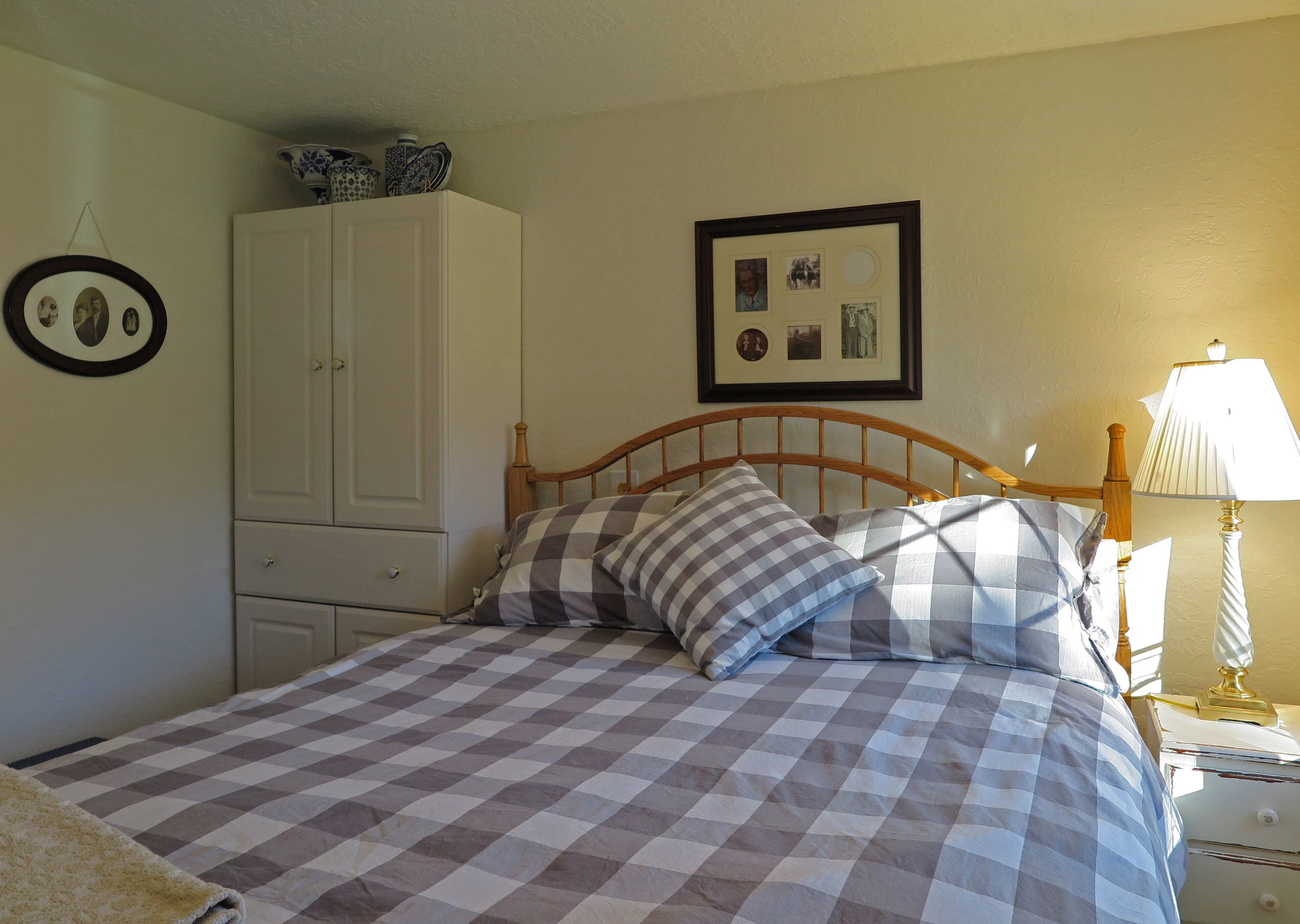 Une chambre avec une couvre-lit à carreaux, une tête de lit en bois, une table de chevet avec une lampe, une armoire et des tableaux encadrés accrochés au mur.