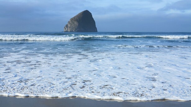 A beach with waves and a large rock formation in the distance. - Gorgeous Ocean View! Private. Cozy Comfort. (Neskowin)