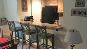A compact kitchen with a refrigerator, a dining area with chairs, and a small table. There is a sign on the wall that reads "SKI MT.SNOW". - Seasons On Mount Snow - Ski-in/Ski-out  (Dover)