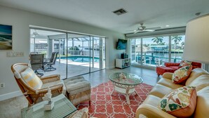 Un vaste salon doté d’une porte vitrée donnant sur un espace piscine, de meubles en osier et d’un tapis rouge à motifs.