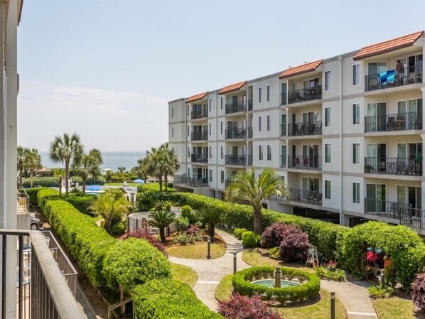 Un hotel de varios pisos con balcones, un jardín ajardinado con una fuente y vistas al mar.