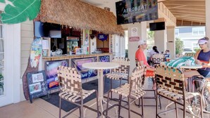 Un espace extérieur aménagé avec des tables et des chaises à toit de chaume, un bar avec une enseigne « Karaoké » et un écran diffusant une sélection des « meilleurs morceaux de 2023 ».