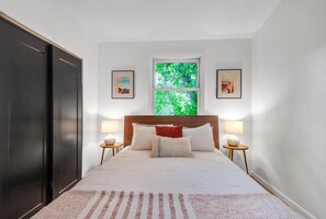A bedroom with a large bed, two bedside tables with lamps, a window with a view of greenery, and two framed pictures on the wall.