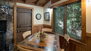 A rustic dining area with a wooden table, chairs, a stone wall, a wooden door, a clock, and a window with a view of trees. - Historic Creekside Cabin with Luxury Amenities, WiFi, Fireplace, Private Trails (Robbinsville)