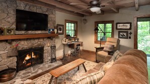 A cozy living room with a stone fireplace, a television mounted on the wall, a comfortable sofa, and a wooden ceiling fan. - Historic Creekside Cabin with Luxury Amenities, WiFi, Fireplace, Private Trails (Robbinsville)