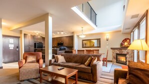 A spacious living area with a sofa, armchair, and coffee table, a kitchen in the background, and a fireplace with a lamp.
