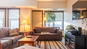 A modern living room with a brown sofa, a wooden coffee table, and a TV on a black cabinet. There are two lamps, a potted plant, and a striped rug.