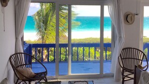 A room with a large sliding glass door leading to a balcony with a view of the ocean. There are two wicker chairs and a small table with a tray.