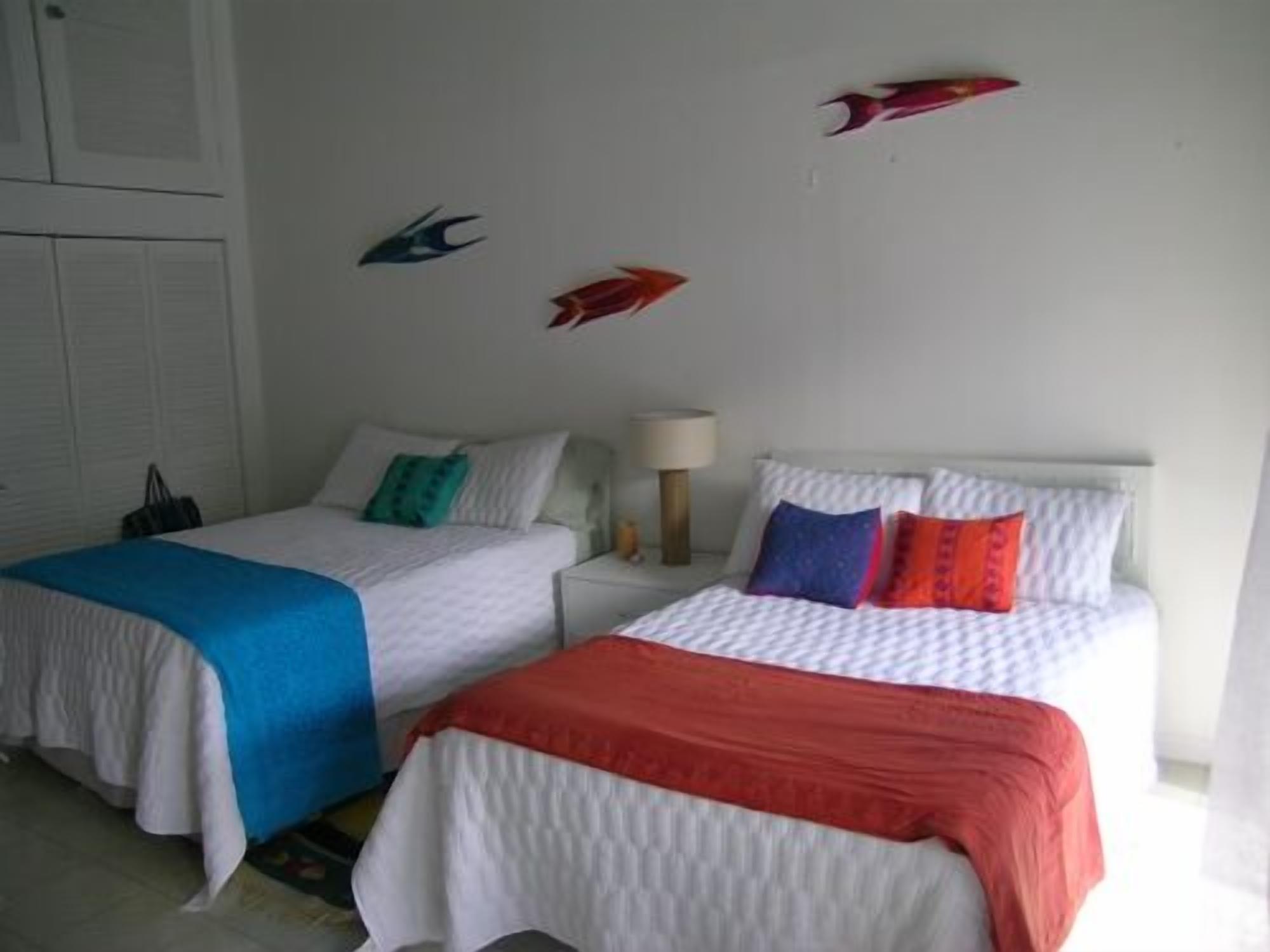 Duas camas de solteiro com roupa de cama branca e almofadas decorativas coloridas, uma mesa de cabeceira com luminária, e uma parede com peixes decorativos.
