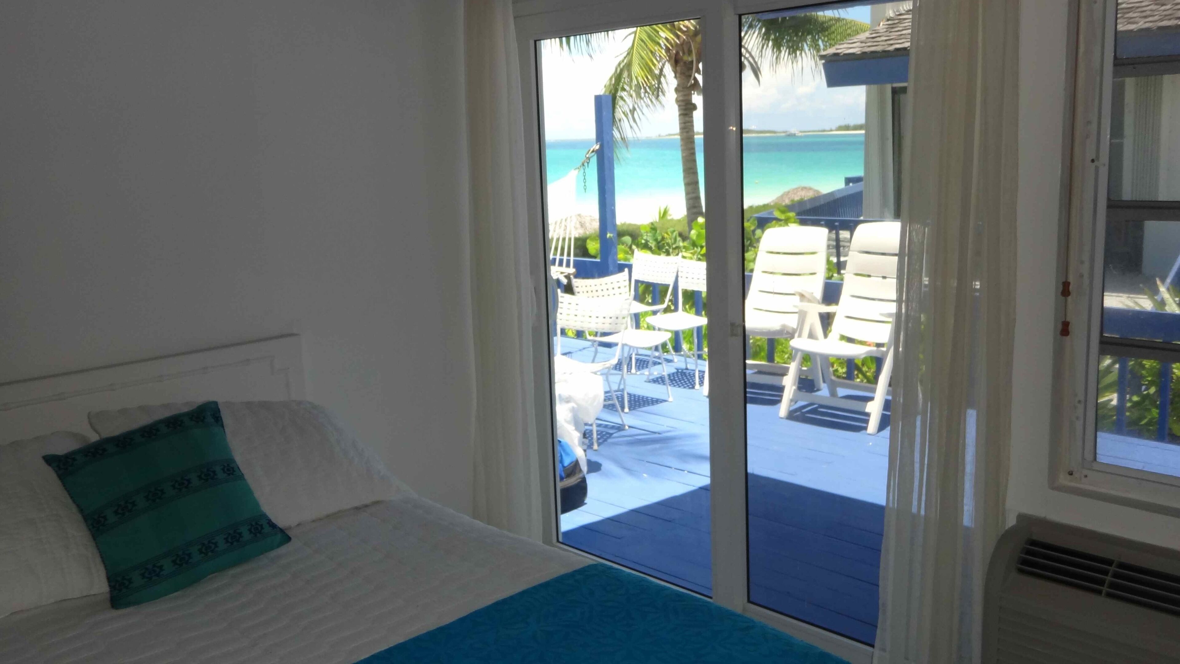 A bedroom with a bed, a view of a balcony with chairs and a palm tree, and a door leading outside.