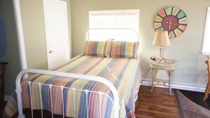 A bedroom with a bed, a bedside table with a lamp, a wall clock, and a window with blinds.