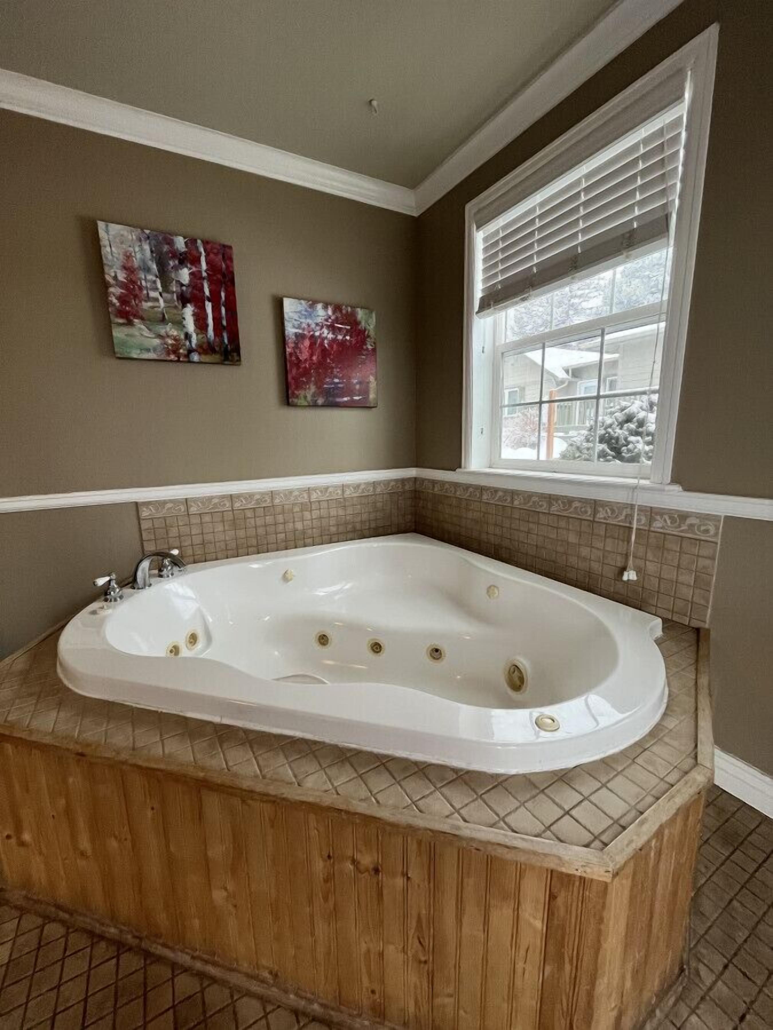 A bathroom with a freestanding bathtub, two wall-mounted artworks, and a window with blinds.
