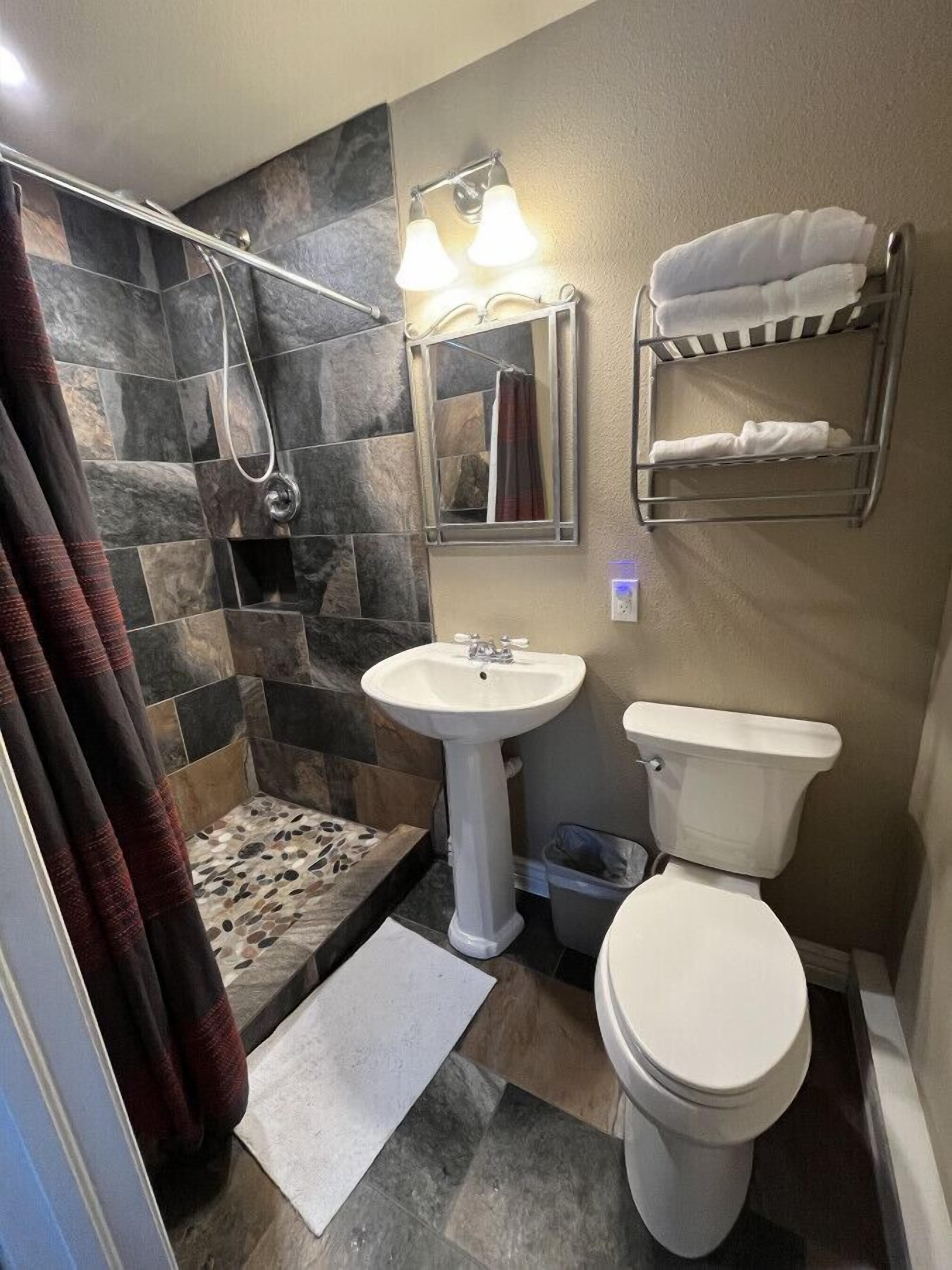 A bathroom with a shower, a sink, and a toilet. There is a towel rack with towels, a mirror, and a window.