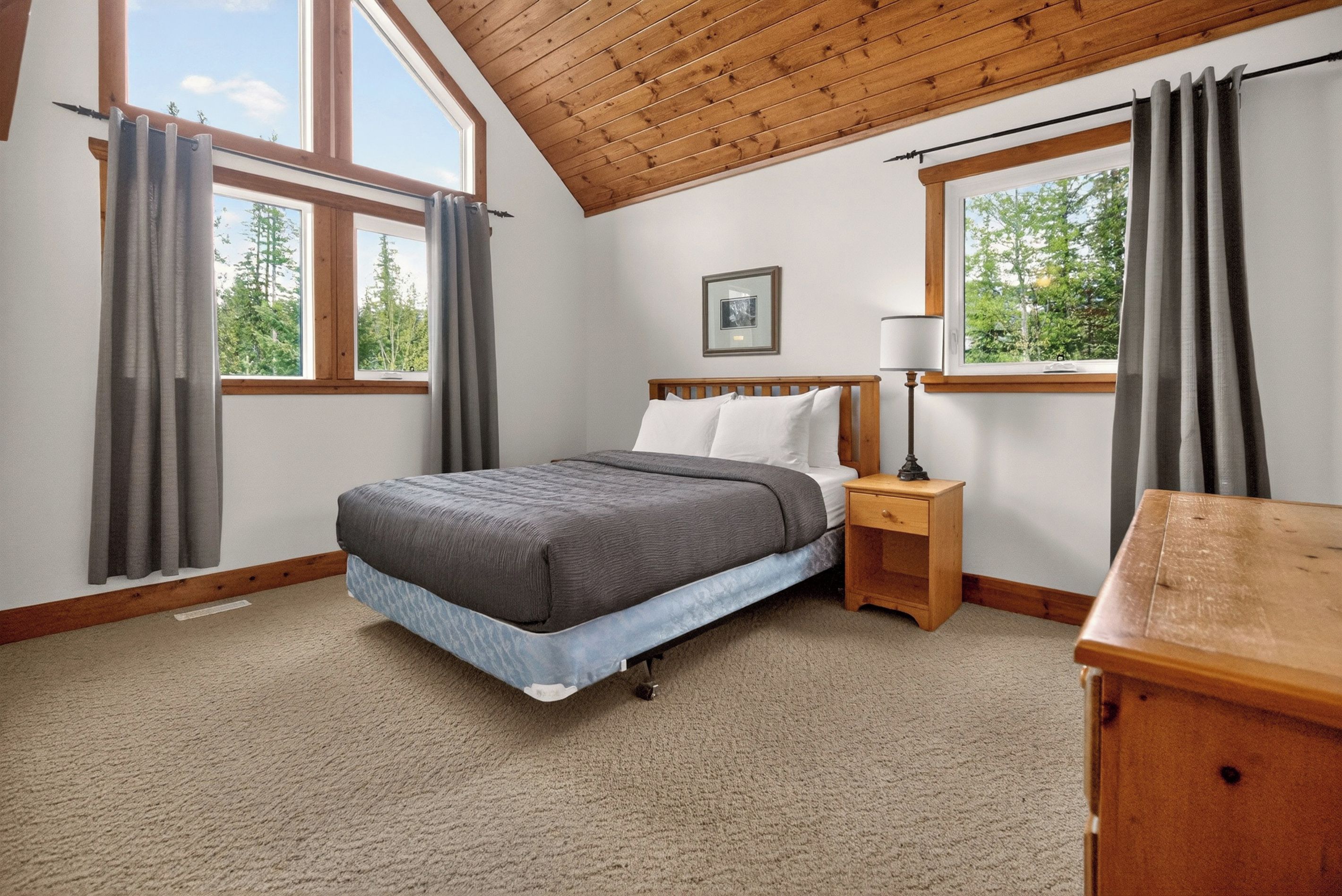 A bedroom with a bed, bedside table, lamp, and a view of trees through the windows.
