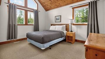 A bedroom with a bed, bedside table, lamp, and a view of trees through the windows.