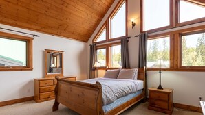 Une chambre avec un lit en bois, une commode, une table de chevet et un miroir. De grandes fenêtres, ornées de rideaux, offrent une vue sur les arbres à l’extérieur.