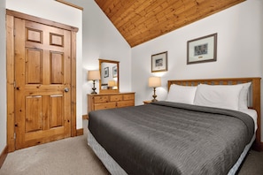 A bedroom with a wooden door, a bed with white bedding, two bedside lamps, a dresser, and two framed pictures on the wall.