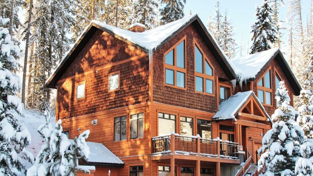 Une grande cabane en bois, dotée de nombreuses fenêtres et d’un balcon, entourée d’arbres couverts de neige.
