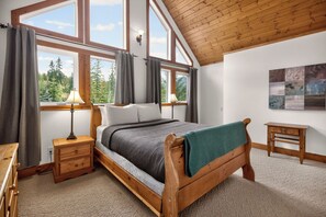 A bedroom with a wooden bed, a nightstand, a lamp, a small table, and a view of trees through large windows.