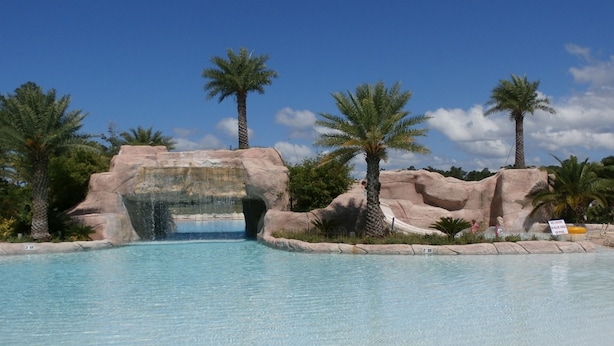 A clear pool with a rock arch entrance, palm trees, and a sign indicating no diving in the blue area.