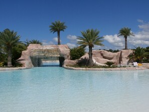 Une piscine aux eaux cristallines, avec une arche rocheuse à l’entrée, des palmiers et un panneau indiquant qu’il est interdit de plonger dans la zone bleue.