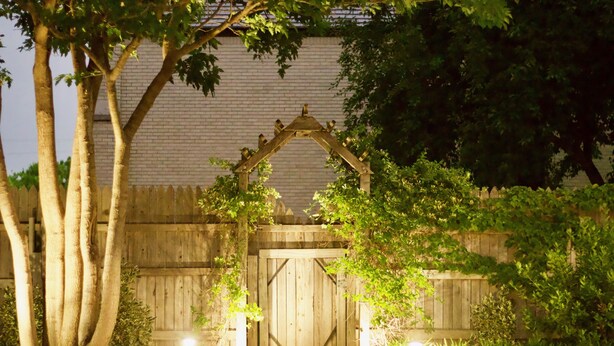 Una reja de madera con un arco decorativo, rodeada de abundante vegetación y un muro de ladrillo.