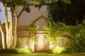Una reja de madera con un arco decorativo, rodeada de abundante vegetación y un muro de ladrillo.