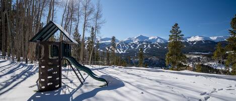 A snow-covered playground with a slide, surrounded by trees and mountains in the background.