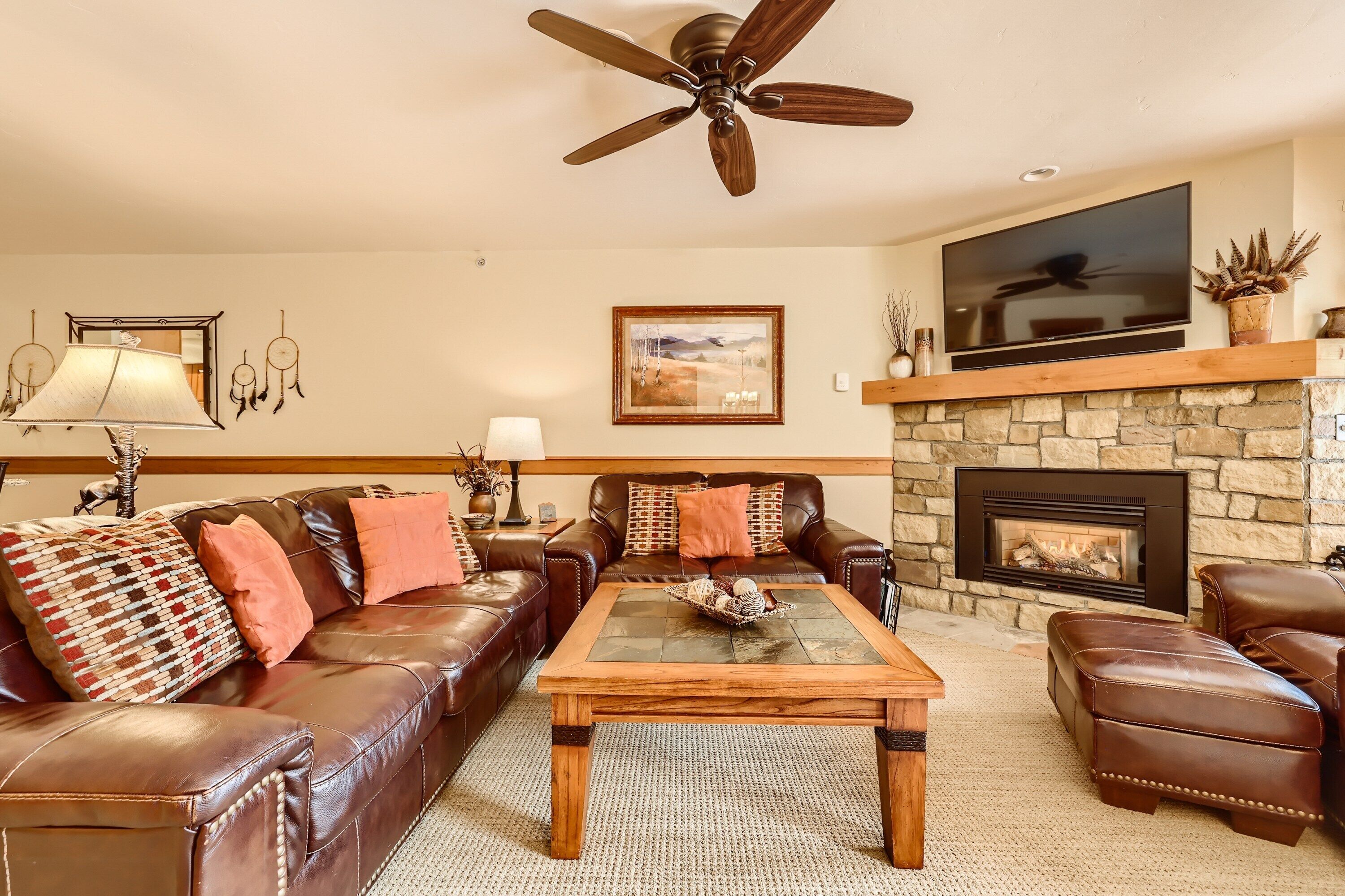 A living room with a brown leather sofa, a wooden coffee table, a stone fireplace, and a ceiling fan.