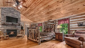 Ein Blockhaushaus-Interieur mit Kamin aus Naturstein, Holzböden, einem Bett mit Blockrahmen, einer Ledersofa und einem Deckenventilator.