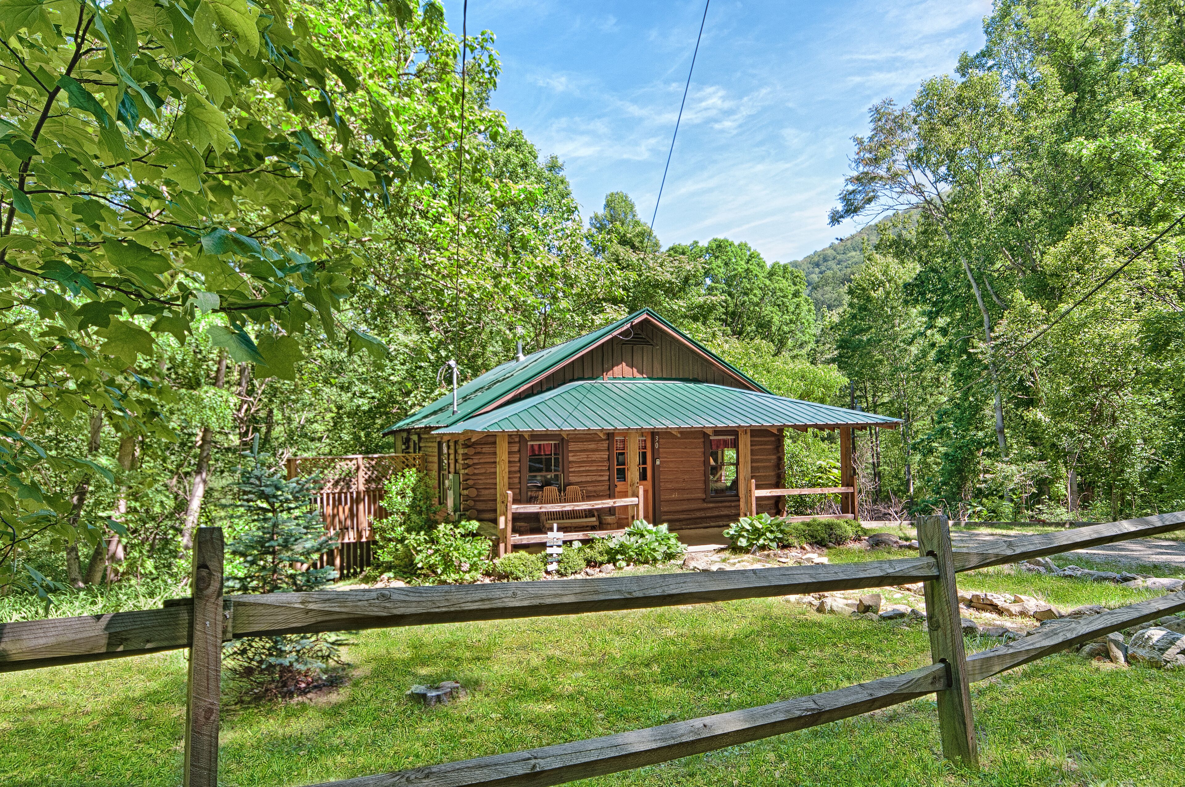 A rustic cabin with a green roof, surrounded by lush greenery and a wooden fence.