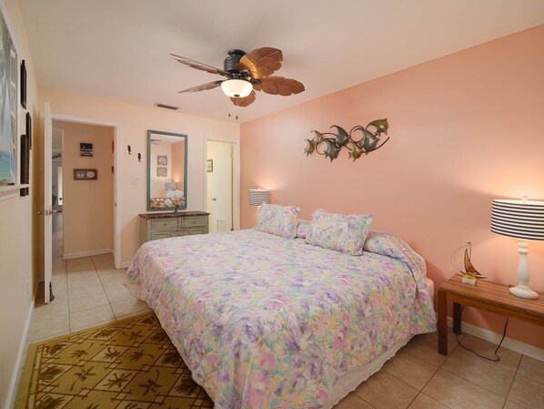 Un dormitorio con una colcha estampada de flores, un ventilador de techo, una lámpara de noche y un adorno en la pared.