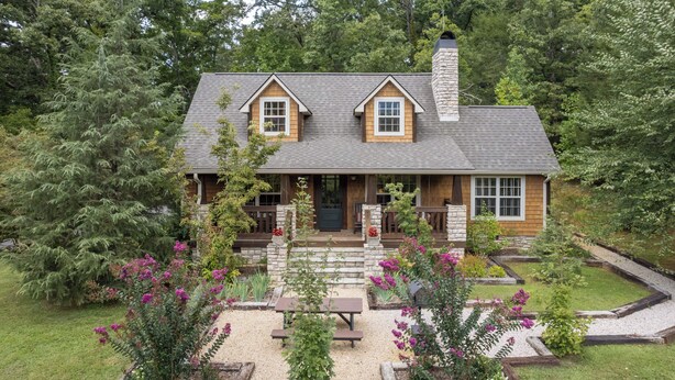 A two-story log cabin with a stone chimney, a front porch with wooden benches, and a garden with flowering plants.