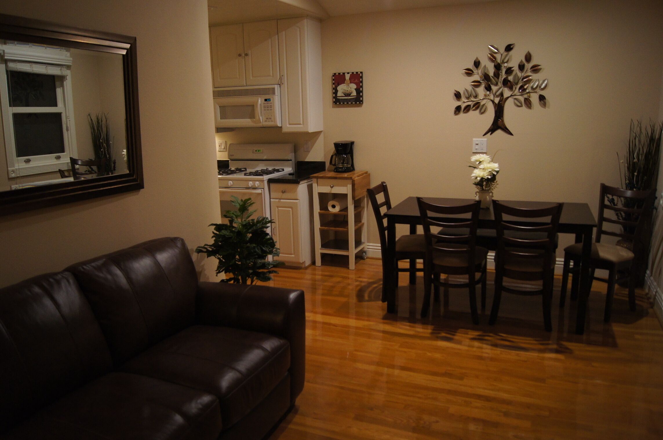 A living room with a brown leather couch, wooden dining table with six chairs, a small potted plant, and a kitchen area with white cabinets and a microwave.
